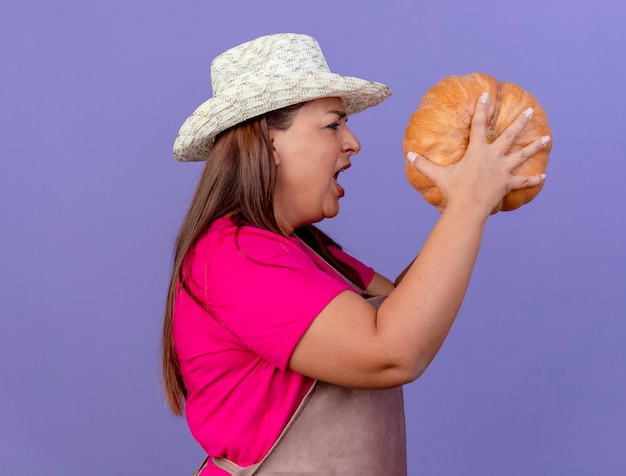 Женщина-садовник средних лет в фартуке и шляпе держит тыкву боком и кричит с сердитым выражением лица на фиолетовом фоне
