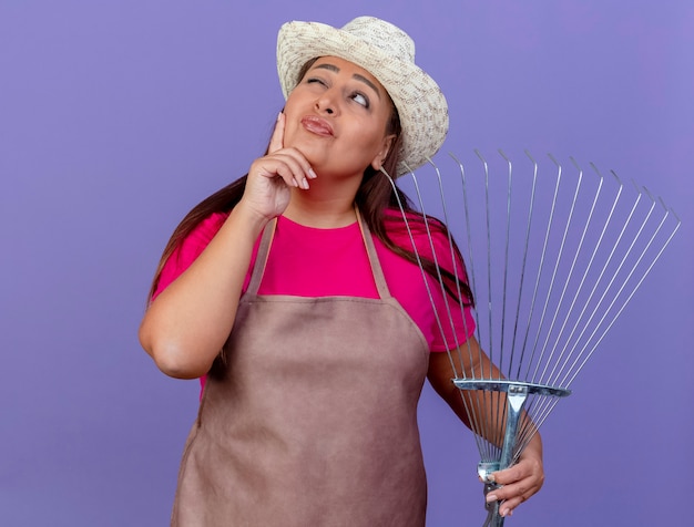 Middle aged gardener woman in apron and hat holding rake looking aside