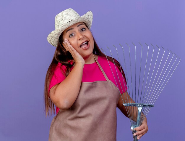 Middle aged gardener woman in apron and hat holding rake smiling being surprised