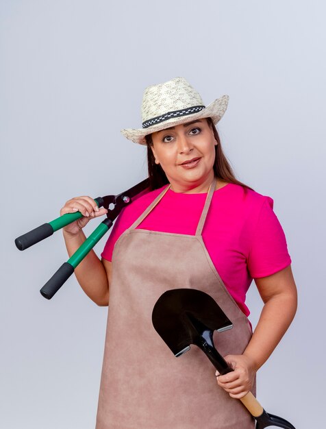 Middle aged gardener woman in apron and hat holding shovel and hedge clippers looking at camera with smile on face standing over white background