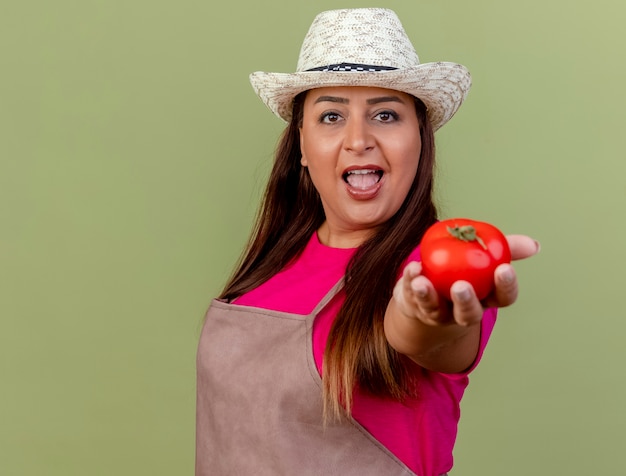 Middle aged gardener woman in apron and hat offering fresh tomato looking at camera smiling standing over light background