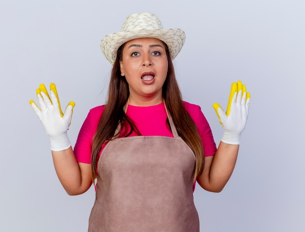 Giardiniere di mezza età donna in grembiule e cappello che indossa guanti di gomma alzando le mani in segno di resa di essere sorpreso e confuso in piedi su sfondo bianco