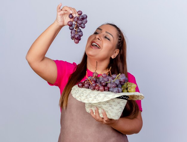 Middle aged gardener woman in apron holding hat full of grapes trying to taste it 