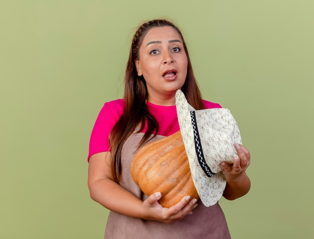 Foto gratuita giardiniere di mezza età donna in grembiule tenendo la zucca con cappello guardando la telecamera confusa in piedi su sfondo chiaro