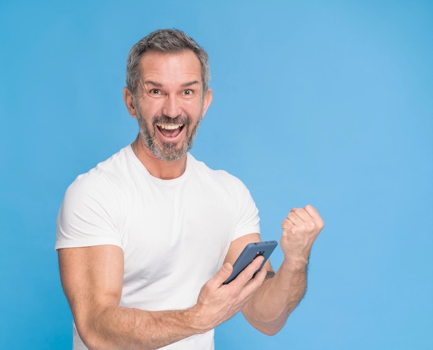 スマートフォンを手に中年の灰色の髪の男は、青い背景に分離された白いtシャツを着てカメラに幸せな笑顔スマートフォンで成熟したフィットの男