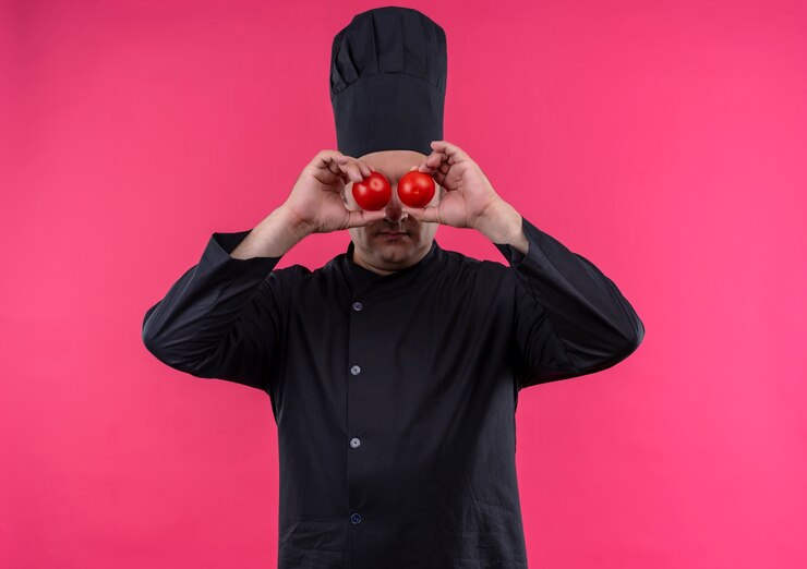 Free Photo middle-aged male cook in chef uniform covered eyes with domates on isolated pink wall