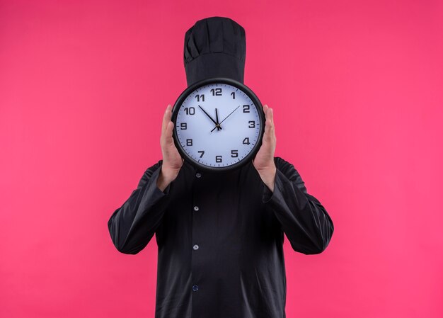 Middle-aged male cook in chef uniform covered face with wall clock on isolated pink wall with copy space