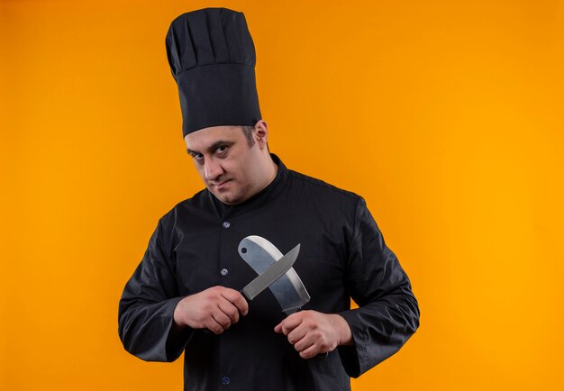 Мужчина-повар средних лет в униформе шеф-повара пересекает тесак и нож на желтой стене с копией пространства
