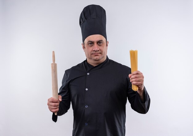 middle-aged male cook in chef uniform holding rolling pin and spaghetti on isolated white wall