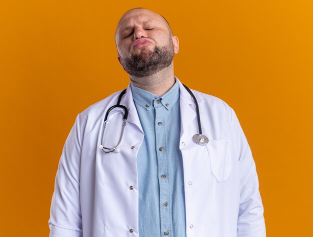 Middle-aged male doctor wearing medical robe and stethoscope doing kiss gesture with closed eyes isolated on orange wall