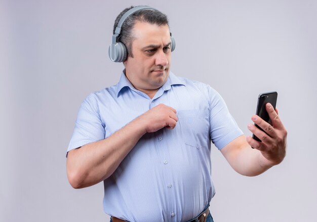 Middle-aged man in blue vertical striped shirt wearing headphones looking at his mobile phone while standing on a white background