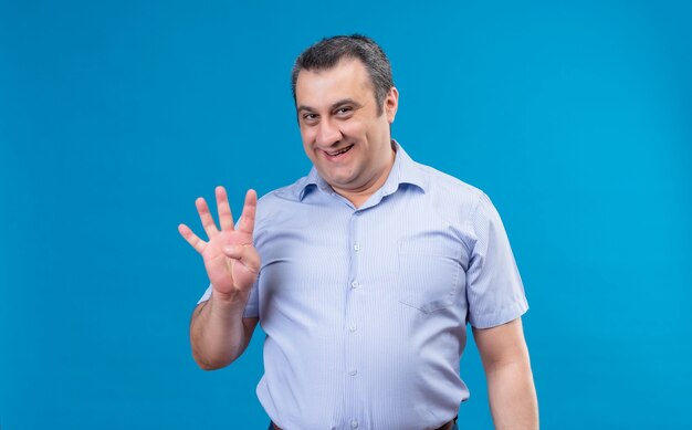 Мужчина средних лет в синей рубашке в вертикальную полоску со счастливым лицом показывает пальцем номер четыре на синем фоне и указывает вверх
