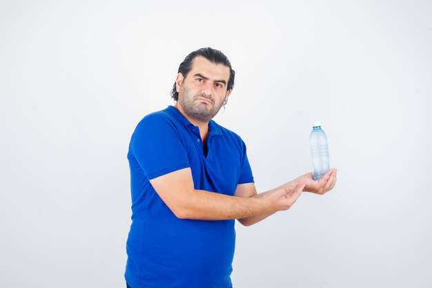 Middle aged man holding water bottle while spreading palm in polo t-shirt and looking bored , front view.