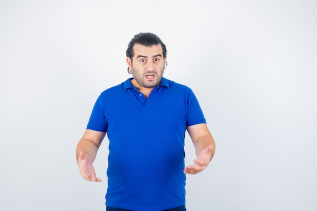 Мужчина средних лет агрессивно держится за руки в синей футболке и выглядит озадаченным