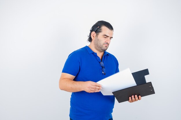 Middle aged man looking over documents in clipboard in polo t-shirt and looking focused , front view.
