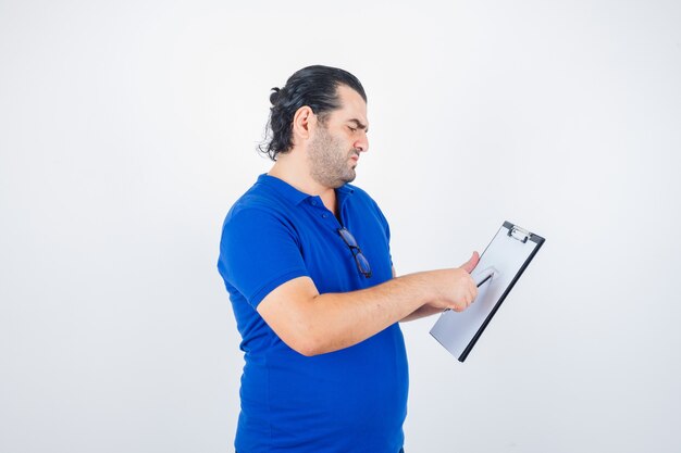 Middle aged man looking through clipboard while holding pencil in polo t-shirt and looking thoughtful , front view.