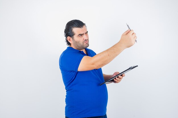 Middle aged man looking through pencil while holding clipboard in polo t-shirt and looking focused. front view.