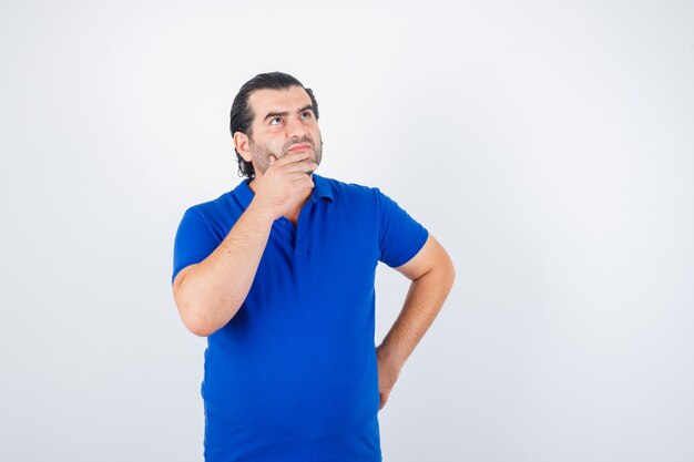 Мужчина средних лет смотрит вверх, держась за бедра и подбородок в синей футболке и выглядит задумчивым