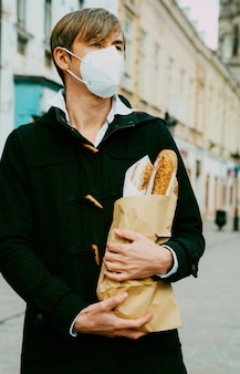 Мужчина средних лет на улице с хлебом, багетом, покупкой буханки во время глобальной пандемии, в маске, достает хлеб из пекарни. еда с собой, покупка булочных во время covid 19.