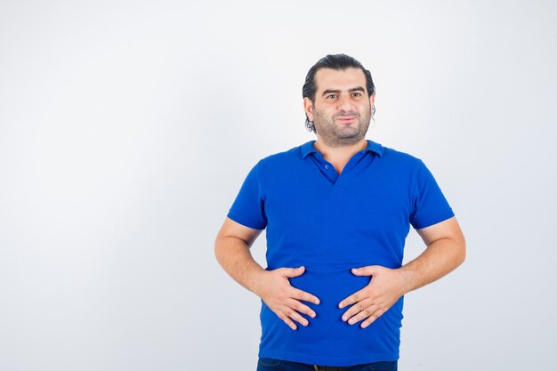 Middle aged man in polo t-shirt keeping hands on stomach and looking joyful