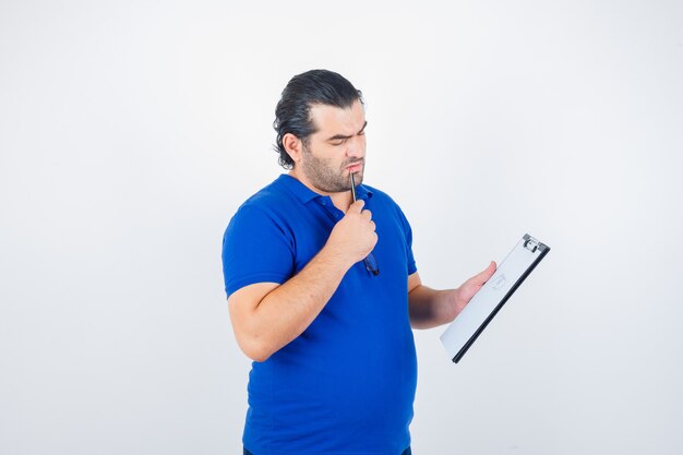 鉛筆を口に持って物思いにふける、正面図を見てクリップボードを通して見ているポロTシャツの中年男性。