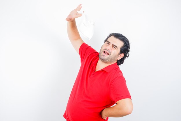 Middle aged man raising napkin while holding hand on hip in red t-shirt and looking thoughtful. front view.