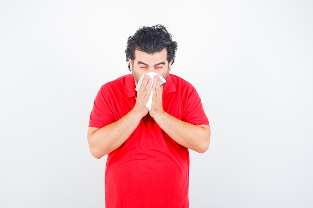 Free photo middle aged man in red t-shirt holding handkerchief blowing running nose and looking unhealthy , front view.