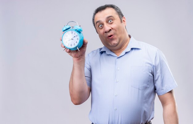 Мужчина средних лет в синей полосатой рубашке держит синий будильник, стоя на белом фоне
