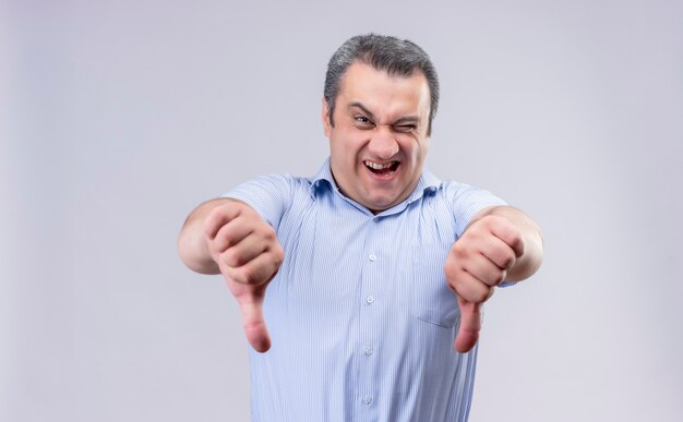 Middle-aged man wearing blue vertical striped shirt showing thumbs down while standing on a white background