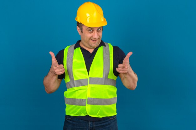 建設黄色いベストと安全ヘルメットを身に着けている孤立した青い壁に笑みを浮かべて人差し指で指している中年の男