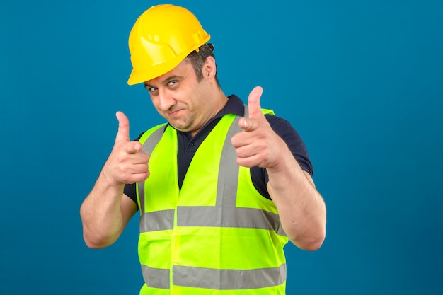 Foto gratuita uomo di mezza età che indossa maglia gialla di costruzione e casco di sicurezza e che puntava il dito indice sorridendo isolato muro blu