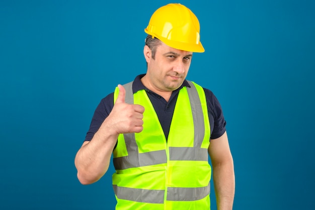 중간 세 남자 건설 노란색 조끼를 입고 웃 고 격리 된 파란색 벽 위에 엄지 손가락을 보여주는 안전 헬멧