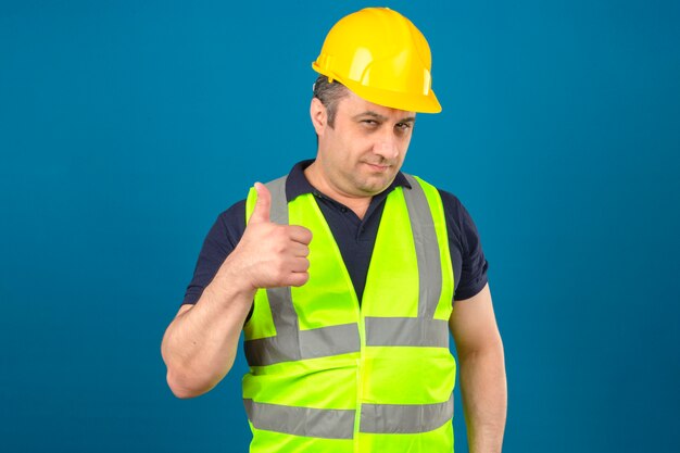 Мужчина средних лет в строительном желтом жилете и защитном шлеме улыбается и показывает палец вверх над изолированной синей стеной