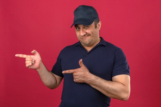 Middle aged man wearing polo shirt and cap pointing with fingers to the side smiling over isolated pink wall