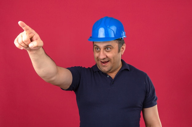 Middle aged man wearing polo shirt and safety helmet pointing to the side with happy face and smiling over isolated pink wall