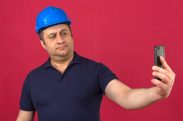Middle aged man wearing polo shirt and safety helmet standing with mobile phone and take a selfie over isolated pink wall