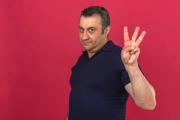 Middle aged man wearing polo shirt with smile on face and showing number three with fingers over isolated pink wall