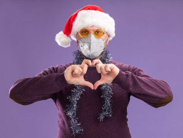 Middle-aged man wearing santa hat and protective mask with tinsel garland around neck with glasses  doing heart sign isolated on purple wall