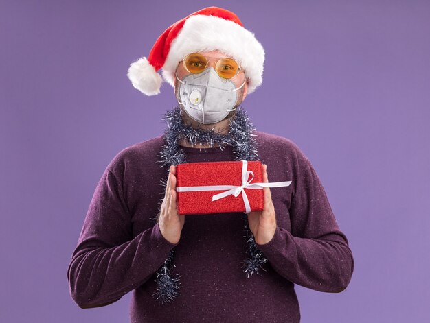 サンタの帽子と保護マスクを身に着けている中年の男性は、紫色の壁に隔離されたギフトパッケージを保持しているメガネと首の周りに見掛け倒しの花輪
