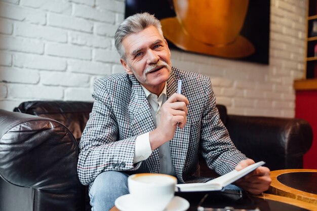 トレンディなスーツを着た中年の年配の男性は、モダンなカフェショップでコーヒーと本を飲みながら素敵な週末を過ごします。