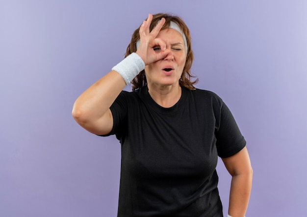 Middle aged sporty woman in black t-shirt with headband doing ok sign looking through this sign surprised standing over blue wall