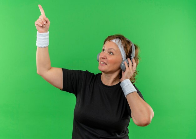 Middle aged sporty woman in black t-shirt with headband and headphones looking aside pointing up with index finger standing over green wall