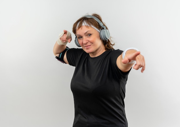 Middle aged sporty woman in black t-shirt with headband and headphones looking confident smiling pointing with index fingers  standing over white wall