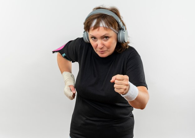 Middle aged sporty woman in black t-shirt with headband and headphones working out looking confident and strained standing over white wall