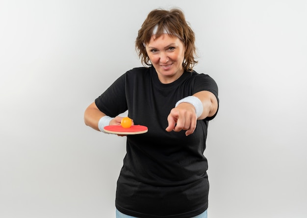 Donna sportiva di mezza età in maglietta nera con fascia tenendo la racchetta con la palla per il ping-pong che punta con il dito alla fotocamera sorridente con la faccia felice in piedi sopra il muro bianco