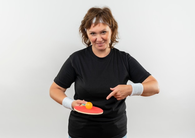 Middle aged sporty woman in black t-shirt with headband holding racket with ball for table tennis pointing with finger to it smiling with happy face standing over white wall