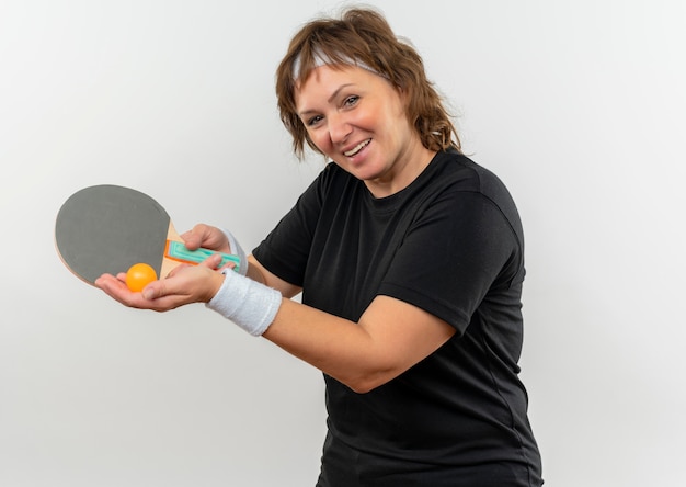 Donna sportiva di mezza età in maglietta nera con fascia tenendo la racchetta con la palla per il ping-pong che punta con sorridente con la faccia felice in piedi sul muro bianco