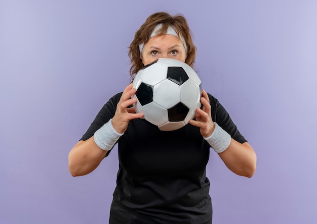 Middle aged sporty woman in black t-shirt with headband holding soccer ball looking confident with serious face standing over blue wall