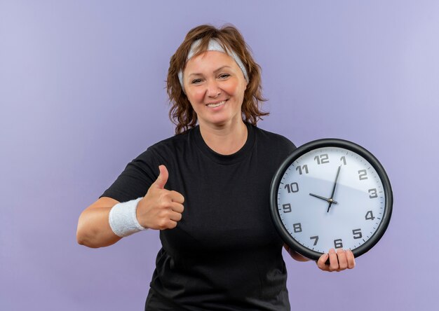 Middle aged sporty woman in black t-shirt with headband holding wall clock smiling cheerfully showing thumbs up standing over blue wall