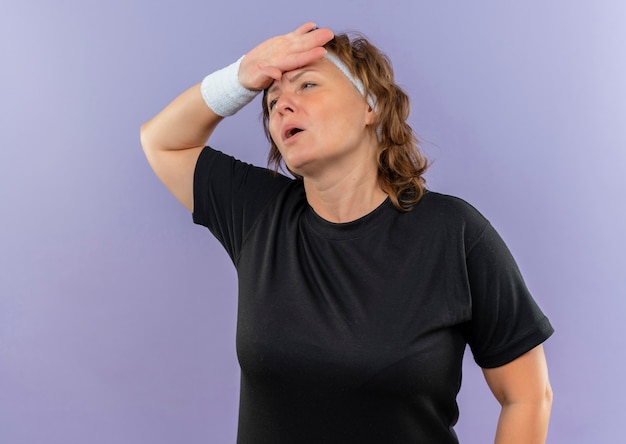 Middle aged sporty woman in black t-shirt with headband looking aside with hand on head tired and exhausted after workout standing over blue wall